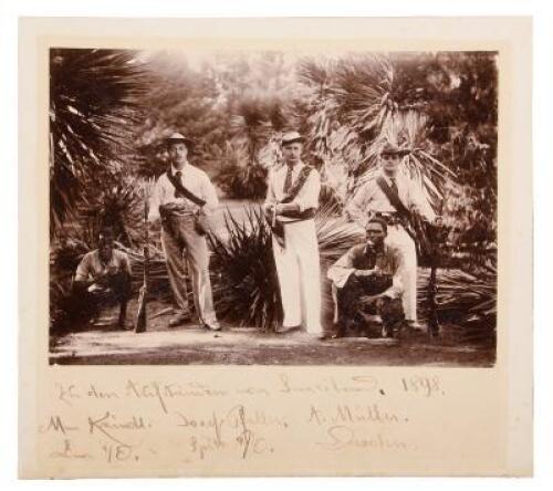 Photograph of three German, or possibly Boer, armed hunters standing in a forest setting with two squatting bearers