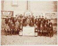 Original sepia-tone photograph of some forty men seated or standing behind a large view of the Pabst Brewery, and two oversize bottles of Pabst beer, and in front of a brick wall on which are hung Pabst banners