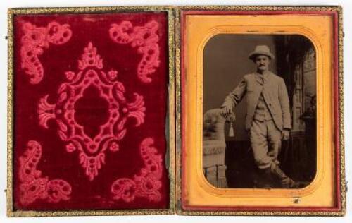Tintype of standing gentleman wearing hat with right hand resting on arm of posing couch