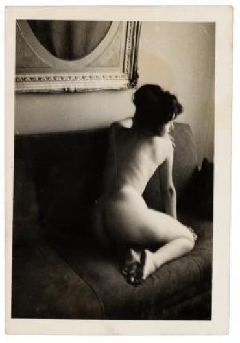 Photograph of a nude woman kneeling on a couch, seen from the back