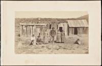 Albumen photograph of two men and four women before a ramshackle hut, two baskets lying on the ground, plus a dog.