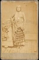 Photograph of young Indian woman in studio, her bare chest covered with beaded necklaces, wearing a beaded skirt