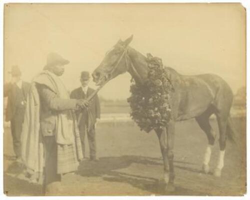 Large original photograph of a thoroughbred racehorse with a large wreath around his neck, identified as Man O' War, being tended by an African American groom, undoubtedly Will Harbut, with two white men in suits observing