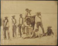 Albumen photograph of 8 Hawaiian boys and young men standing or seated by or on a horse at the beach, with straw hats and leis