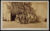 Albumen photograph of two men, four women and a baby beneath a tree in front of a warehouse or barn, with some form of agricultural press